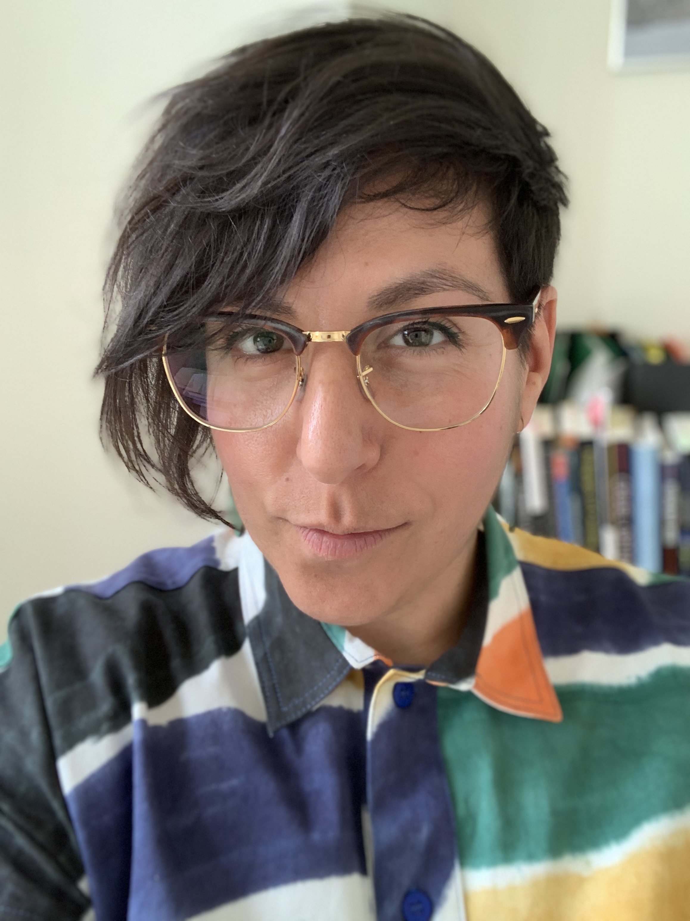 Photograph of Natalie Kouri-Towe; person wearing glasses with asymmetrical brown hair, and a blue, green and black colour blocked shirt.